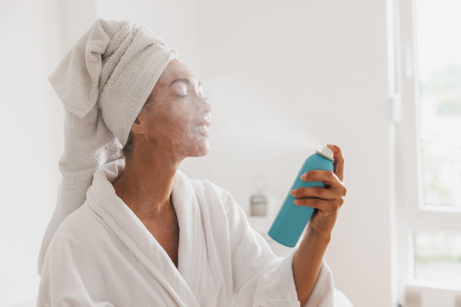 Woman in a white robe and hair towel spraying face mist on her skin