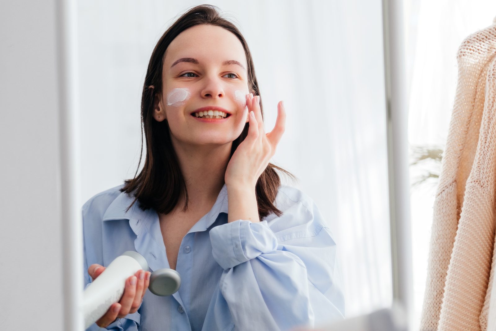 Woman smoothing cream onto her face at the bathroom mirror in the morning