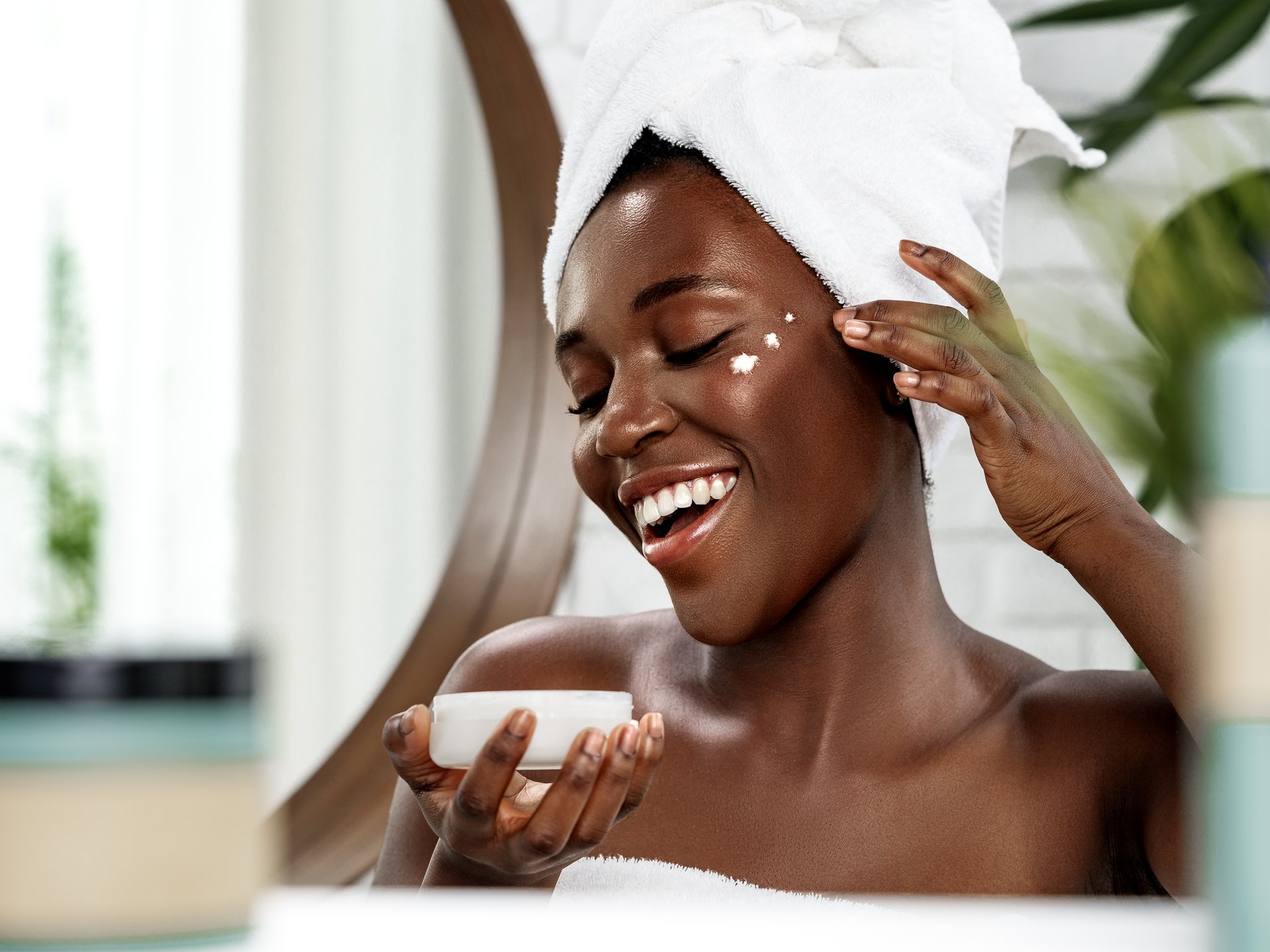 Smiling woman with her hair in a towel dabbing face cream onto her cheek, holding the open jar