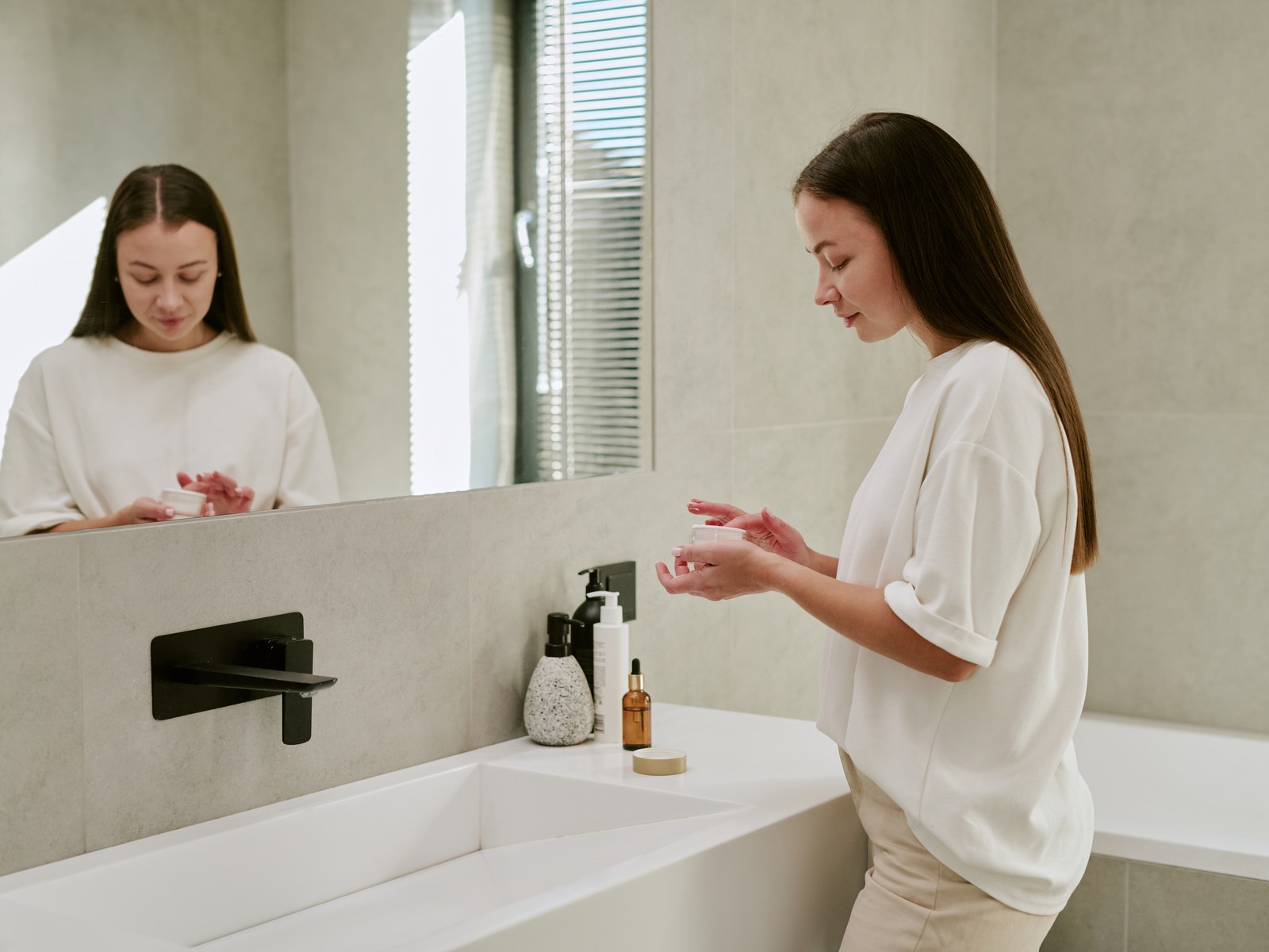 Woman applying moisturiser at a bright bathroom sink beside a large mirror